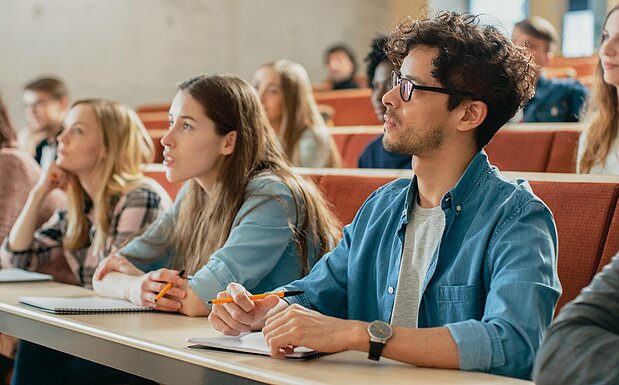Mehrere Studierende sitzen in einem Hörsaal und hören einem Dozenten zu. 